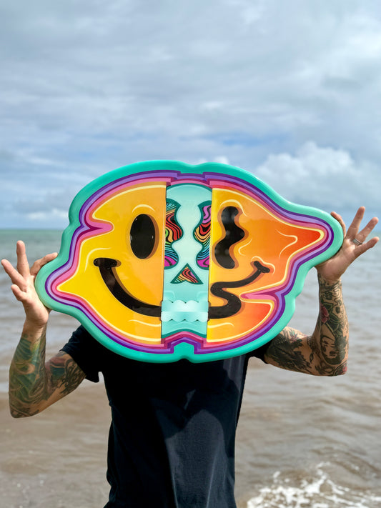 Person holding a colorful brain-shaped object with a smiley face design over their face at the beach.