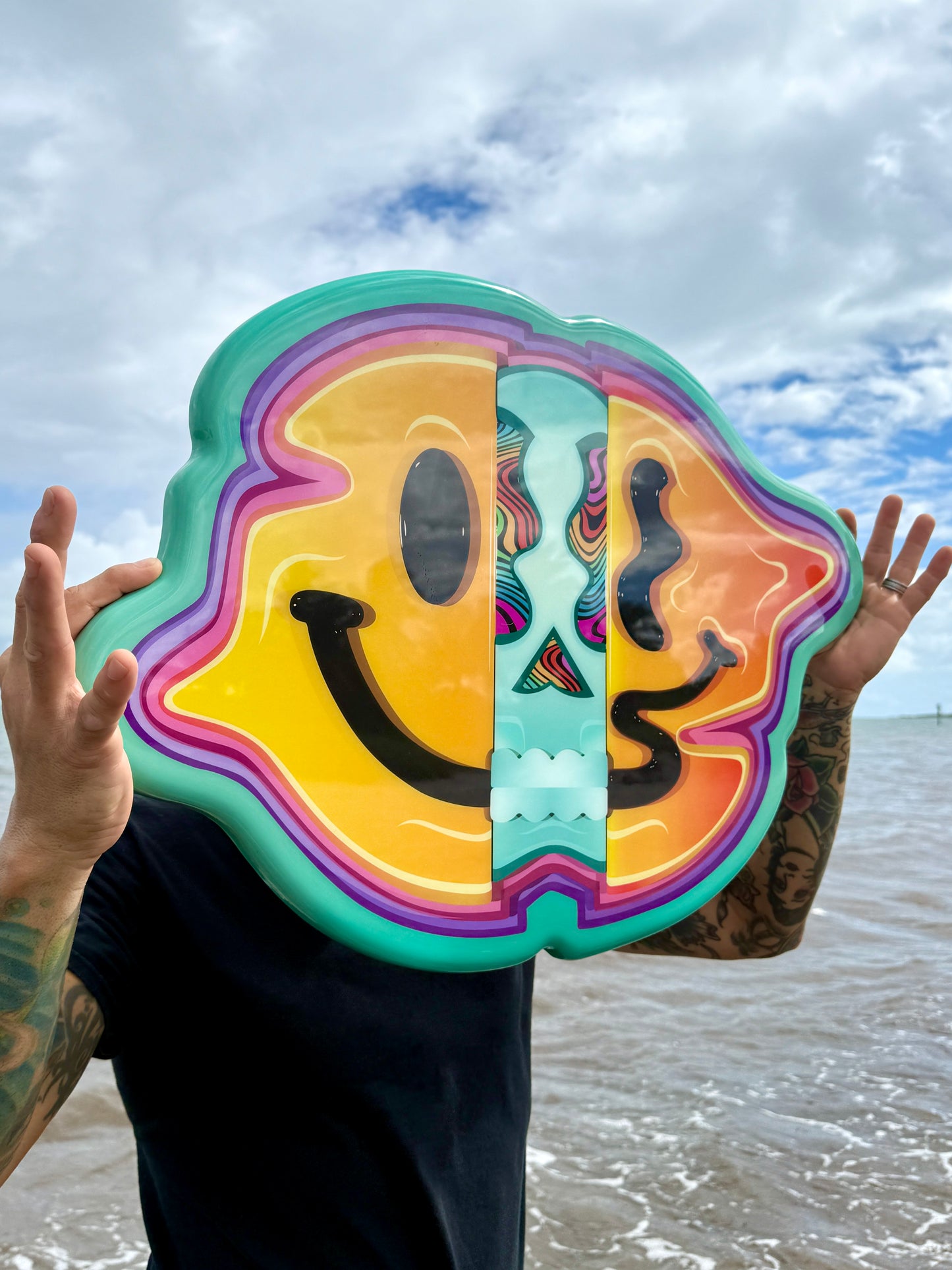 Person holding a colorful smiley face object with a beach in the background